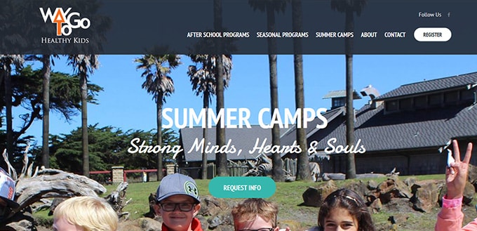 Children smiling outdoors at a summer camp with trees and a building in the background.